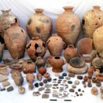 Fig. 27. The assemblage from Pillared Hall (L.1040) in Palace B at Khirbet al-Batrawy (Zarqa), which includes pithoi, red-burnished jugs and pointed juglets, medium-sized jars, ceremonial vessels with applied decorations of snakes and scorpions, and noteworthy objects, among which are a bone knife and several worked bones, pierced seashells, and a carefully worked basalt potter's wheel (L. Nigro).