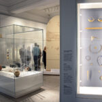 Fig. 2. A view in the Penn Museum’s Middle East Galleries. On the right, finely crafted jewelry made of gold and precious stones, excavated from the royal tombs at Ur. The central case features materials from the excavation of a warrior’s grave, dating to ca. 100 years after the royal tombs at Ur, likely the tomb of an important warrior, who was richly adorned and buried along with numerous weapons. In the background is visible the resin-cast reconstruction of Ur-Namma’s stele from ca. 2100 B.C.E. (courtesy Penn Museum, Philadelphia).